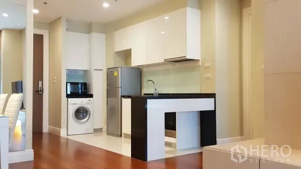 Condominium for rent in Khlong Toei, Bangkok - Modern open-plan kitchen with white cabinets, black countertop island, fridge and washing machine on wood flooring.