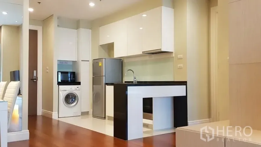 Condominium for rent in Khlong Toei, Bangkok - Modern open-plan kitchen with white cabinets, black countertop island, fridge and washing machine on wood flooring.