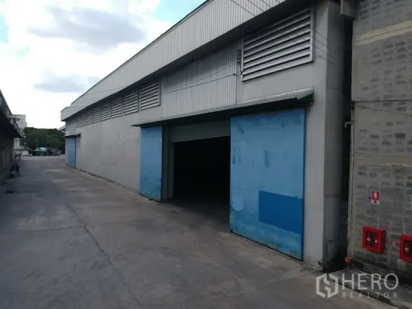 Factory for rent in Phra Pradaeng, Samut Prakan - Large industrial warehouse exterior with blue sliding doors, ventilation louvres, and a wide truck-friendly driveway.