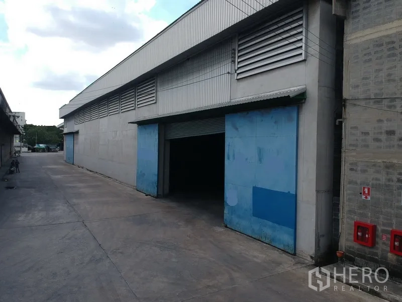 Factory for rent in Phra Pradaeng, Samut Prakan - Large industrial warehouse exterior with blue sliding doors, ventilation louvres, and a wide truck-friendly driveway.