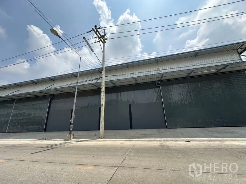 Warehouse for rent in Lam Luk Ka, Pathum Thani - Front elevation of the long warehouse facade with multiple large sliding doors along the main road.