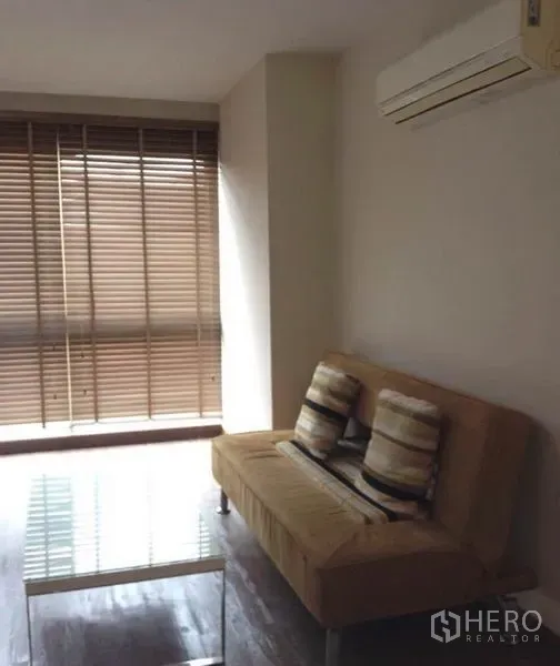 Condominium for rent in Khlong Toei, Bangkok - Cozy living area with tan sofa, air conditioner, glass coffee table and large window blinds.