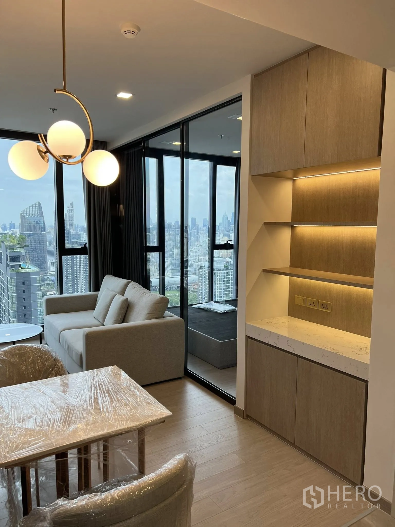 คอนโด for rent in Huai Khwang, Bangkok - ชั้นบิลท์อินพร้อมไฟส่องและโซฟา ข้างผนังกั้นกระจกของห้องนอน มองออกสู่วิวเมืองกว้าง