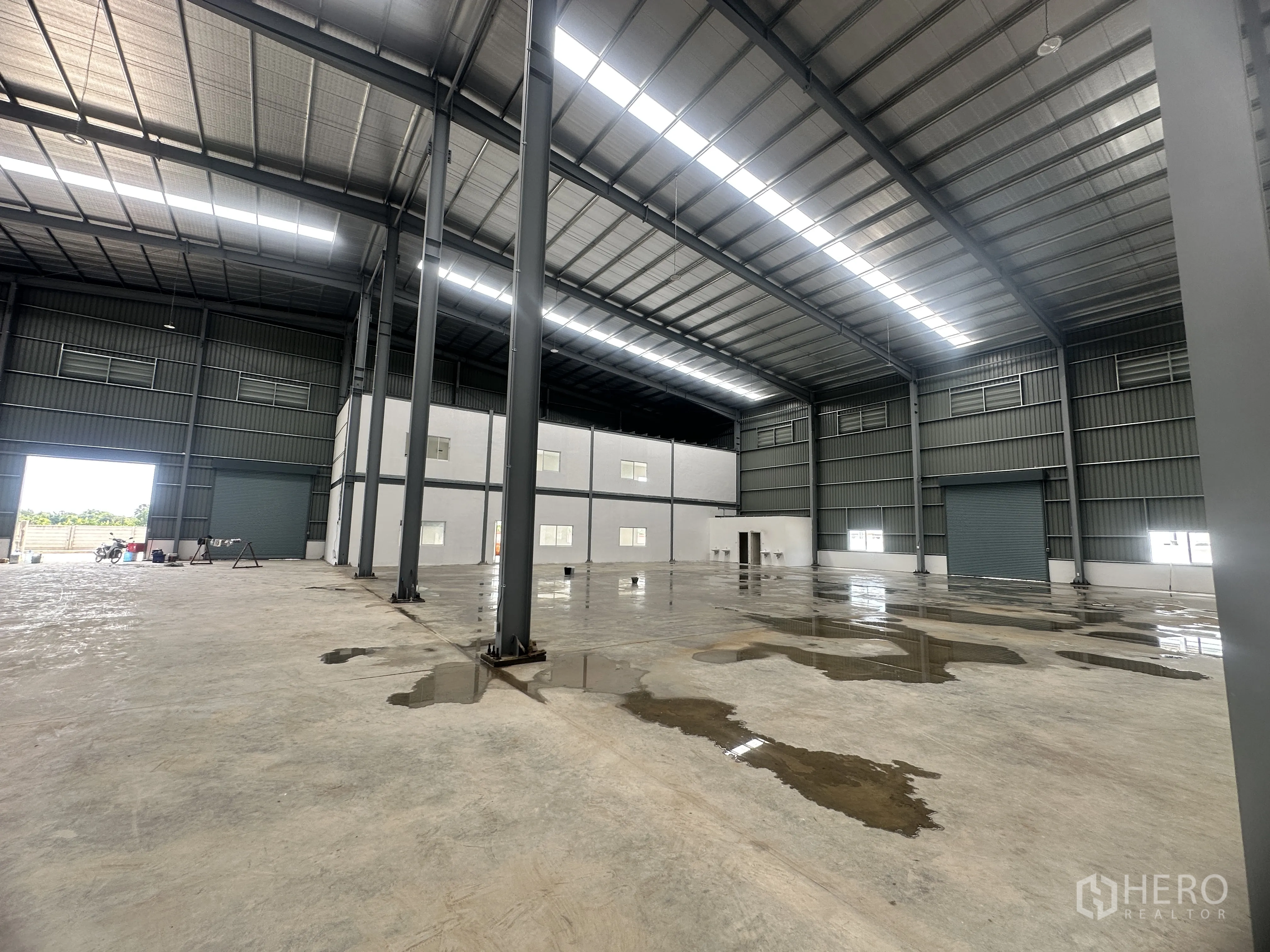 Warehouse for rent or sale in Lam Luk Ka, Pathum Thani - Warehouse interior with an office block, several roller shutters, and wet concrete floor patches.