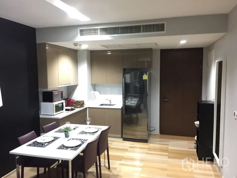 Condominium for rent in Khlong Toei, Bangkok - Modern L-shaped kitchen with glossy cabinets, four-seat dining table, and stainless-steel refrigerator.