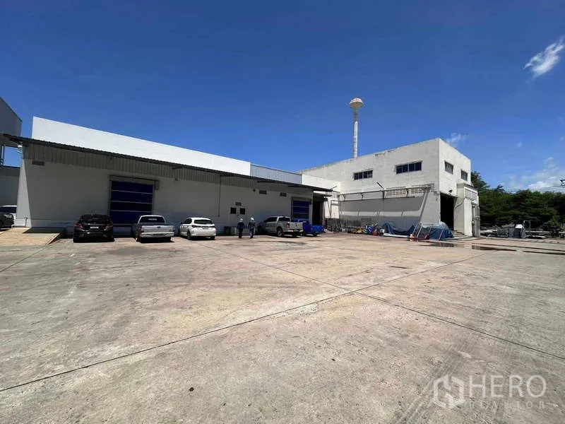 โรงงาน ให้เช่า - อาคารโรงงานภายนอก ประตูม้วนสีน้ำเงิน ลานคอนกรีตกว้าง และที่จอดรถใต้อากาศแจ่มใส