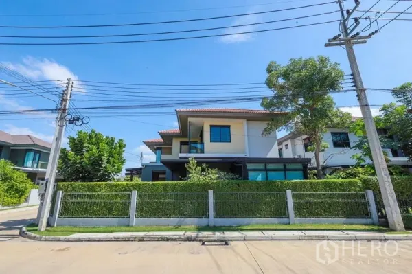 Detached House for rent or sale in Lam Luk Ka, Pathum Thani - Street view of the contemporary corner house behind a trimmed hedge fence.