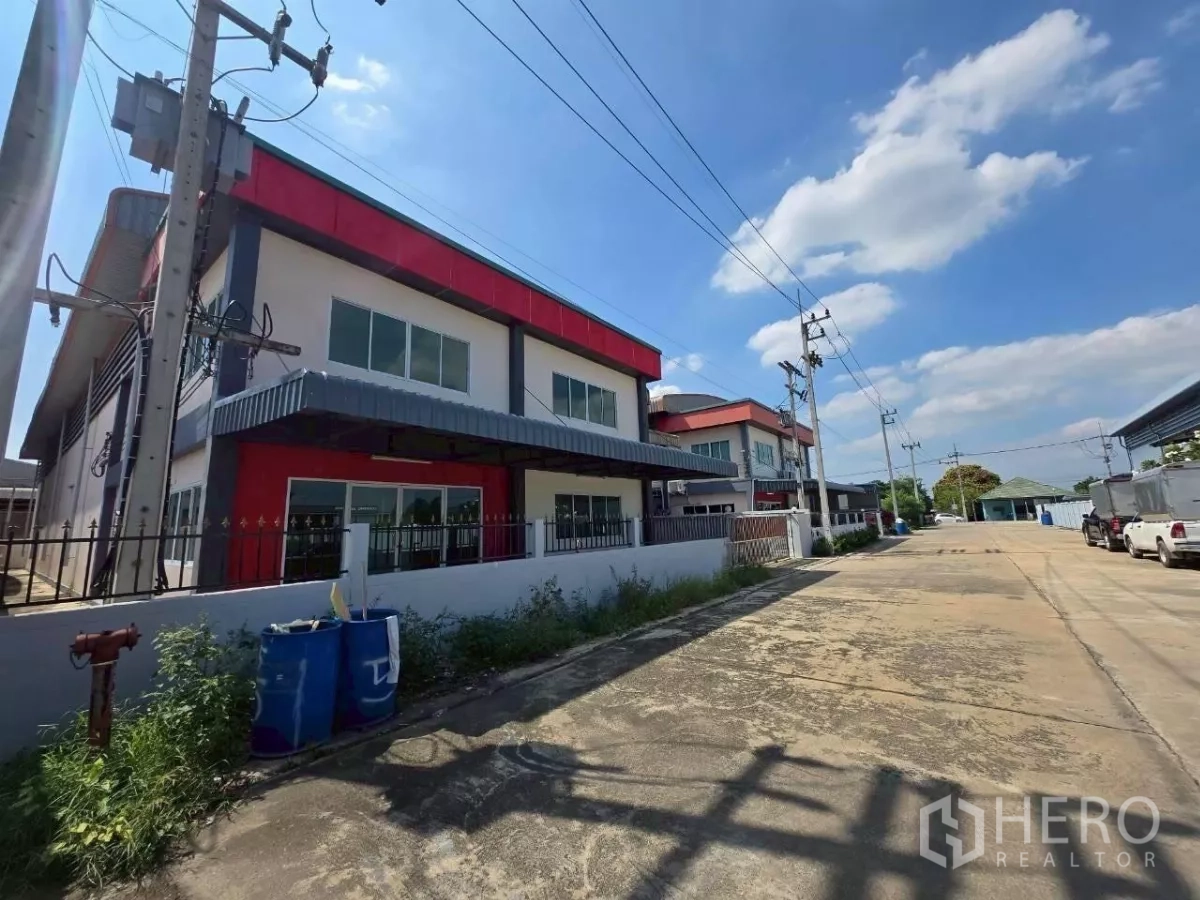 Factory for rent or sale in Mueang Samut Sakhon, Samut Sakhon - Street view of a modern two-storey factory with red accents, fenced perimeter and a wide access road in Samut Sakhon.