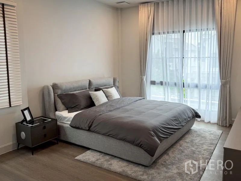 Detached House for rent or sale - Sunlit bedroom with a gray upholstered bed, large windows, and an area rug.