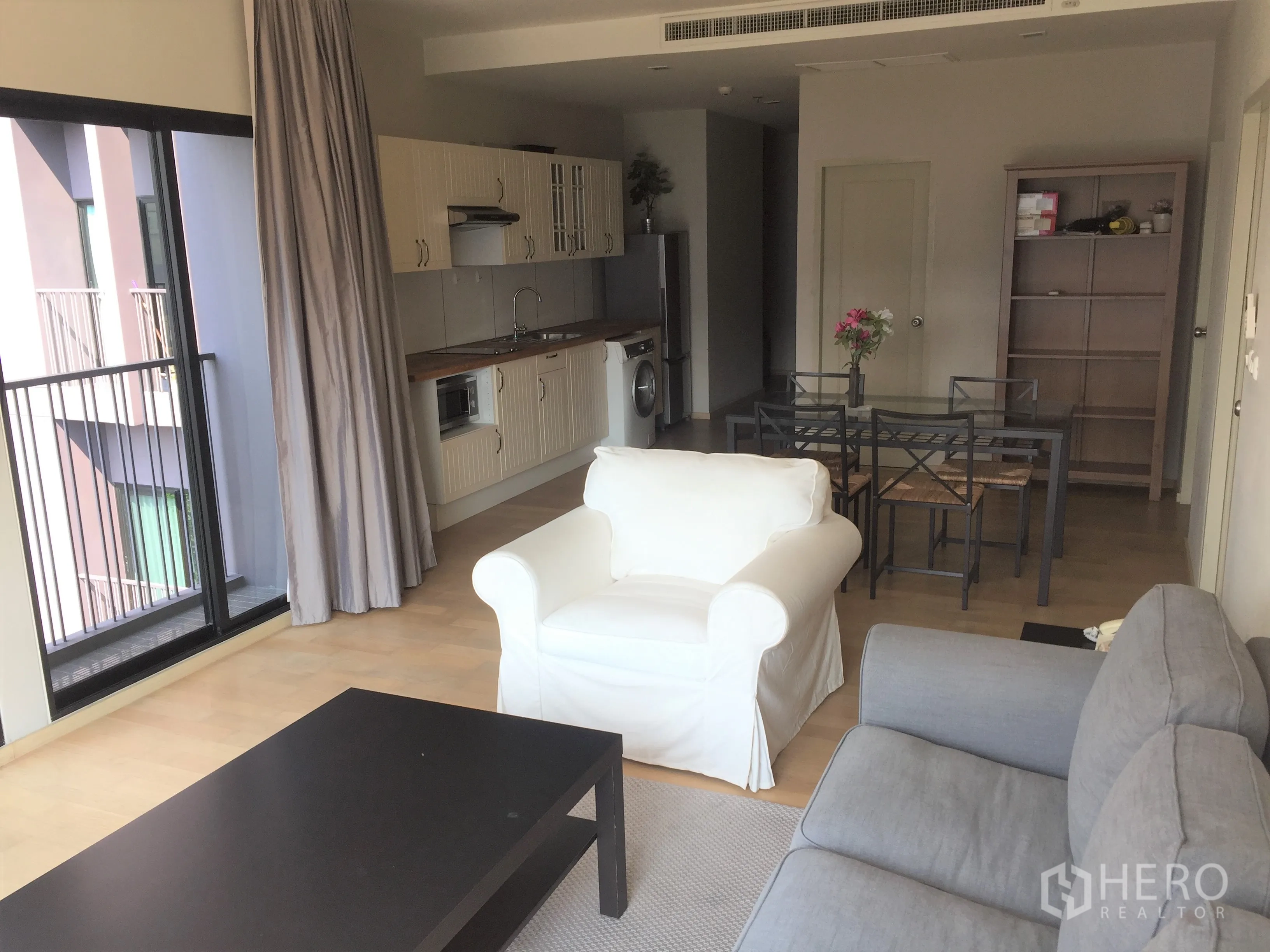 Condominium for rent or sale in Watthana, Bangkok - Bright open-plan living room with balcony doors, light wood floors, sofas and a fitted kitchenette.