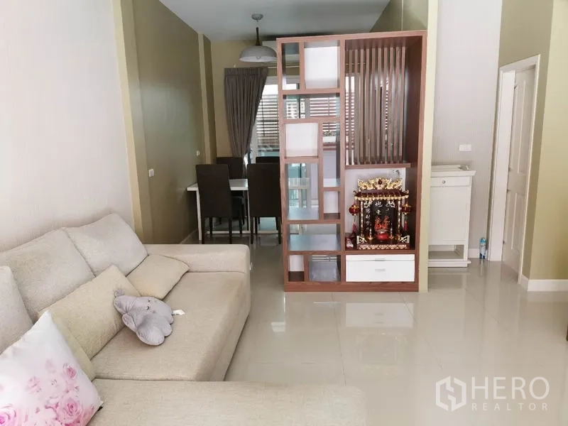 联排别墅 出租在Wang Thonglang, Bangkok - 明亮客厅，L 型沙发与亮面瓷砖地面，装饰隔断通向餐区。