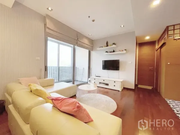 Condominium for sale in Khlong Toei, Bangkok - Bright living room with cream sectional sofa, wall-mounted TV and sliding doors to a balcony.