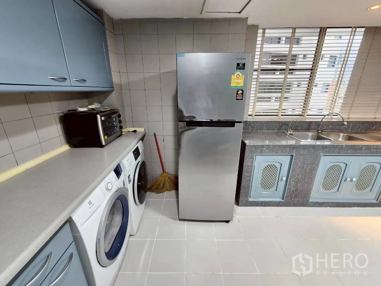 Condominium for rent in Khlong Toei, Bangkok - Kitchen corner with refrigerator, washer and dryer beside a double sink and blinds.