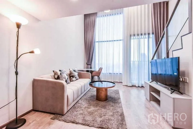 Condominium for rent in Huai Khwang, Bangkok - Light-filled lounge with sofa, rug and tall windows beside staircase.