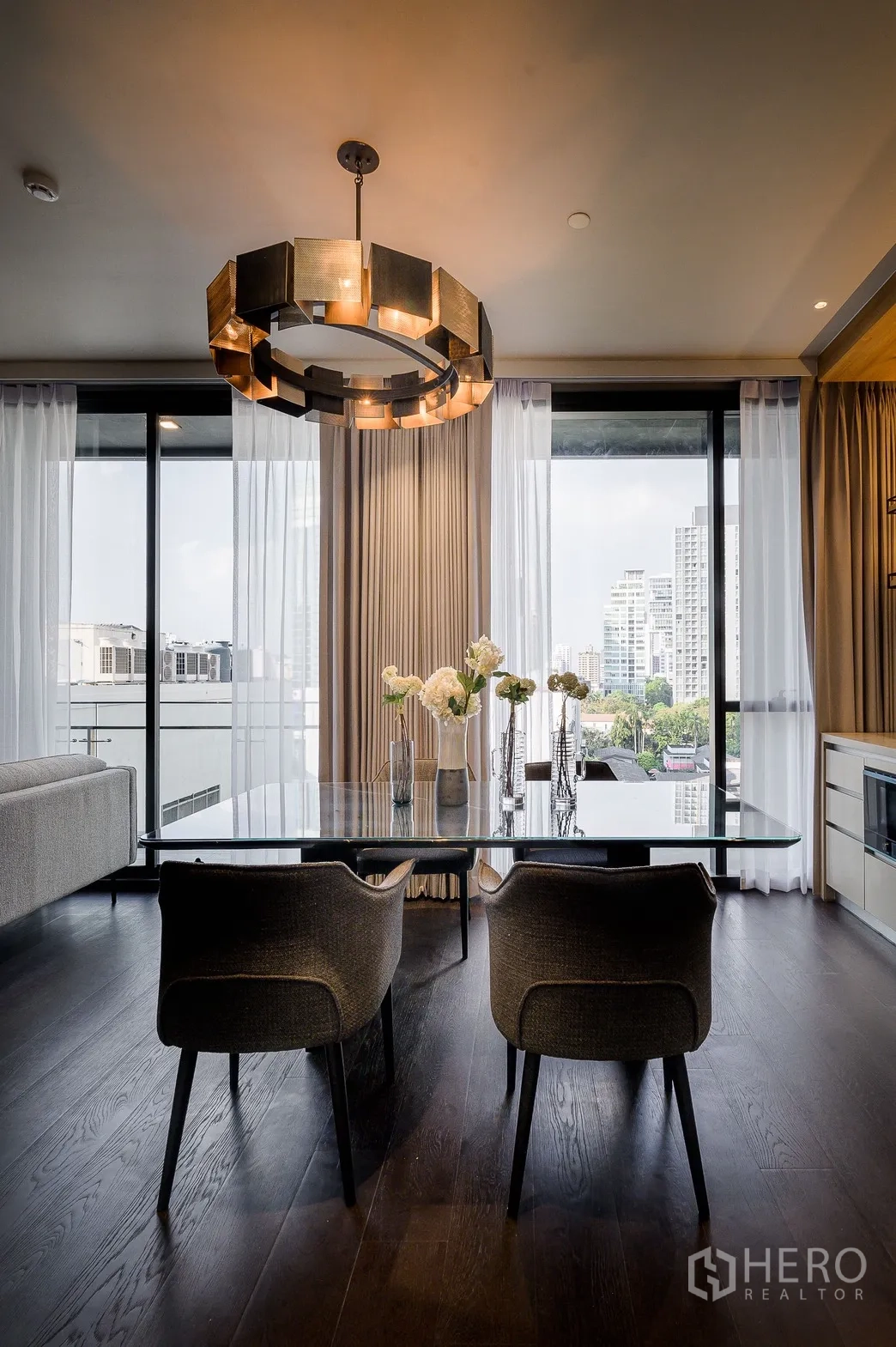 Condominium for rent or sale in Watthana, Bangkok - Dining space with glass table, floral centerpiece and sculptural ring chandelier beside large windows.