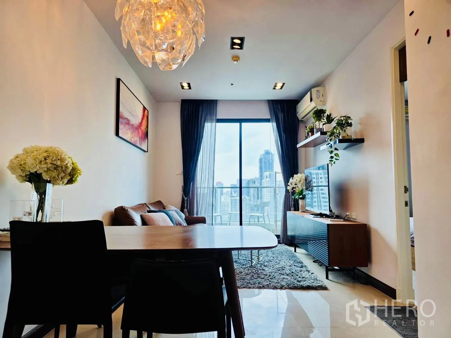 คอนโด for rent in Huai Khwang, Bangkok - โต๊ะรับประทานอาหารและโซฟาใต้โคมไฟดีไซน์ ข้างประตูกระจกสูงจรดเพดานออกสู่ระเบียงชมเมือง