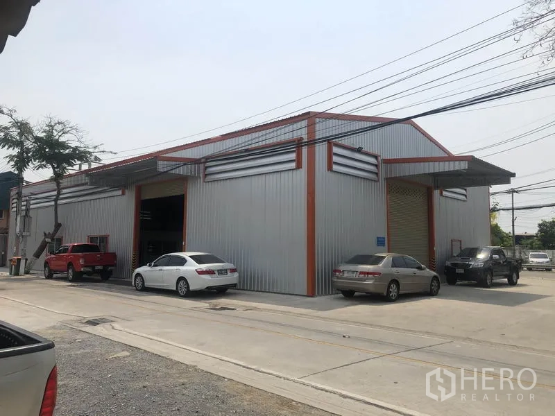 Warehouse for rent in Bang Yai, Nonthaburi - Corner view of a modern corrugated-steel warehouse with cars parked along a wide concrete street.