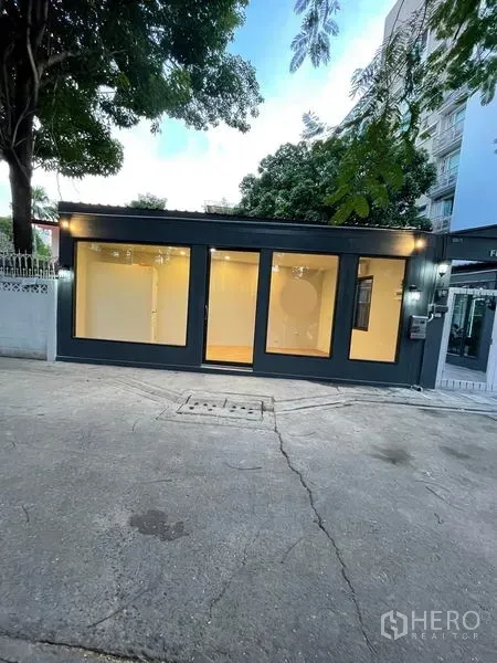 零售空间 出租在Khlong Toei, Bangkok - 临街单层零售铺面，深色外框与整面落地玻璃，室内暖色灯光，周围有树与公寓，位于曼谷空堤区。