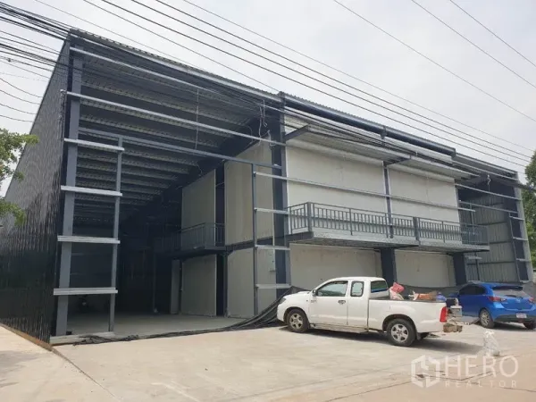 Warehouse for rent in Lam Luk Ka, Pathum Thani - Wide front view of twin-bay warehouse facade with balconies and parked vehicles.