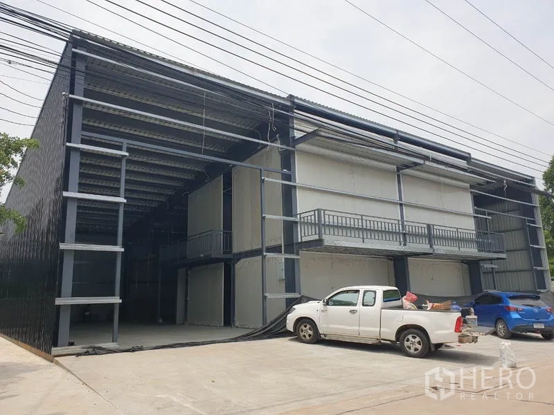 Warehouse for rent in Lam Luk Ka, Pathum Thani - Wide front view of twin-bay warehouse facade with balconies and parked vehicles.