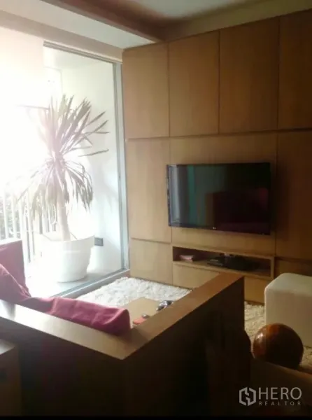 公寓 出租在Watthana, Bangkok - 客厅配内嵌电视墙、木质沙发与长绒地毯，推门通往带绿植的阳台。