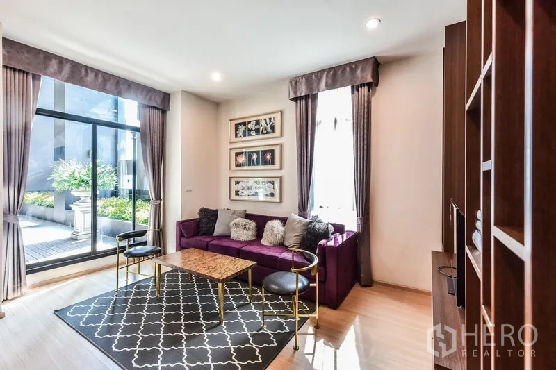 Condominium for rent or sale in Huai Khwang, Bangkok - Cozy living area with floor-to-ceiling windows, purple sectional sofa, patterned rug, and access to a plant-filled terrace.