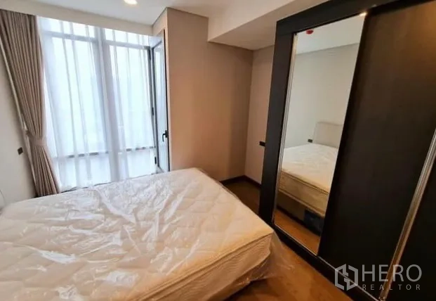 Condominium for rent in Khlong Toei, Bangkok - Bedroom with double bed beside tall windows and mirrored wardrobe.