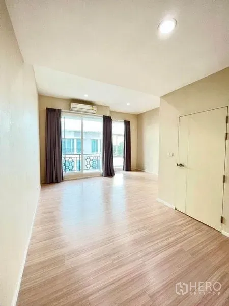 Townhouse for sale in Bang Kapi, Bangkok - Bright room with wood-look flooring, sliding glass doors to a balcony, air conditioner, and dark curtains.