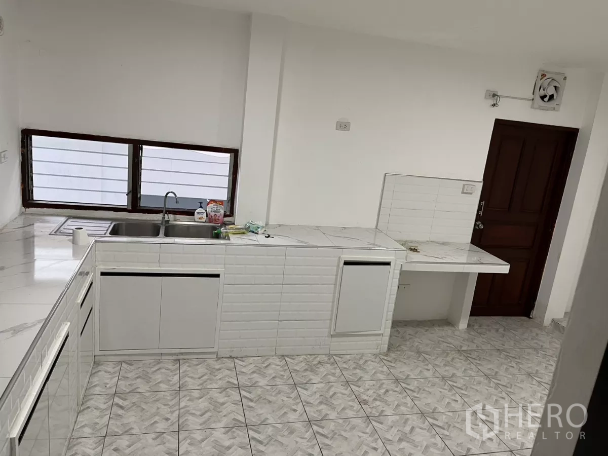 Detached House for rent in Watthana, Bangkok - Bright kitchen with white cabinetry, marble-look counters, double sink and tiled floor.