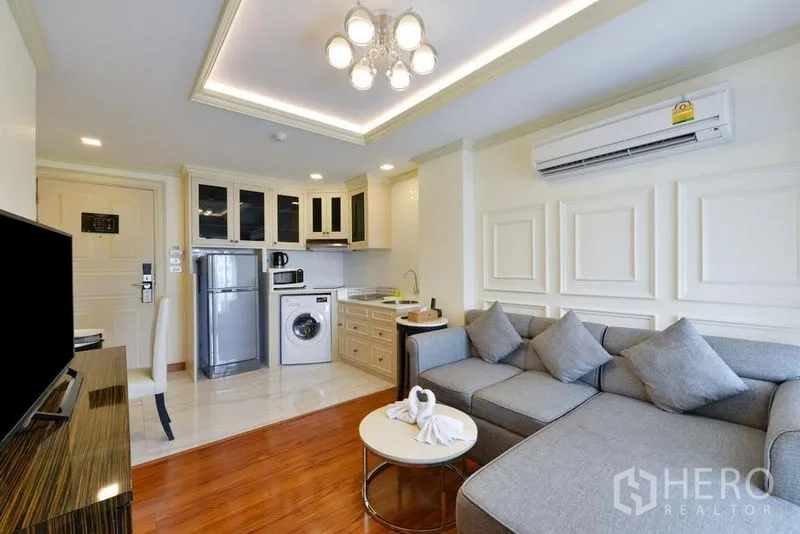 Apartment for rent in Khlong Toei, Bangkok - Open-plan living area with L-shaped sofa, ceiling light, air-con, and kitchenette.