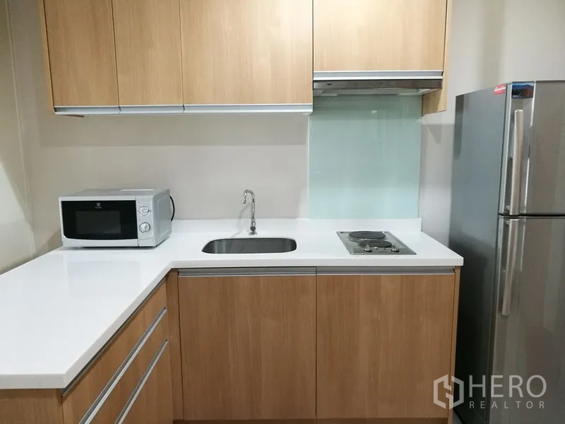 Condominium for rent in Ratchathewi, Bangkok - Compact U‑shaped kitchen with wood cabinets, white countertop, cooktop, hood, microwave and fridge.