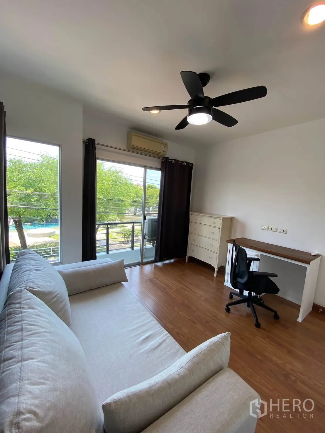 独栋房屋 for rent in Mueang Samut Prakan, Samut Prakan - 采光充足的房间，配沙发与书桌，阳台望向绿地与泳池。