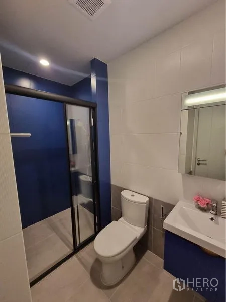 公寓 出租在Watthana, Bangkok - 现代卫浴，蓝色点缀墙，玻璃淋浴间、白色洗手台与马桶。