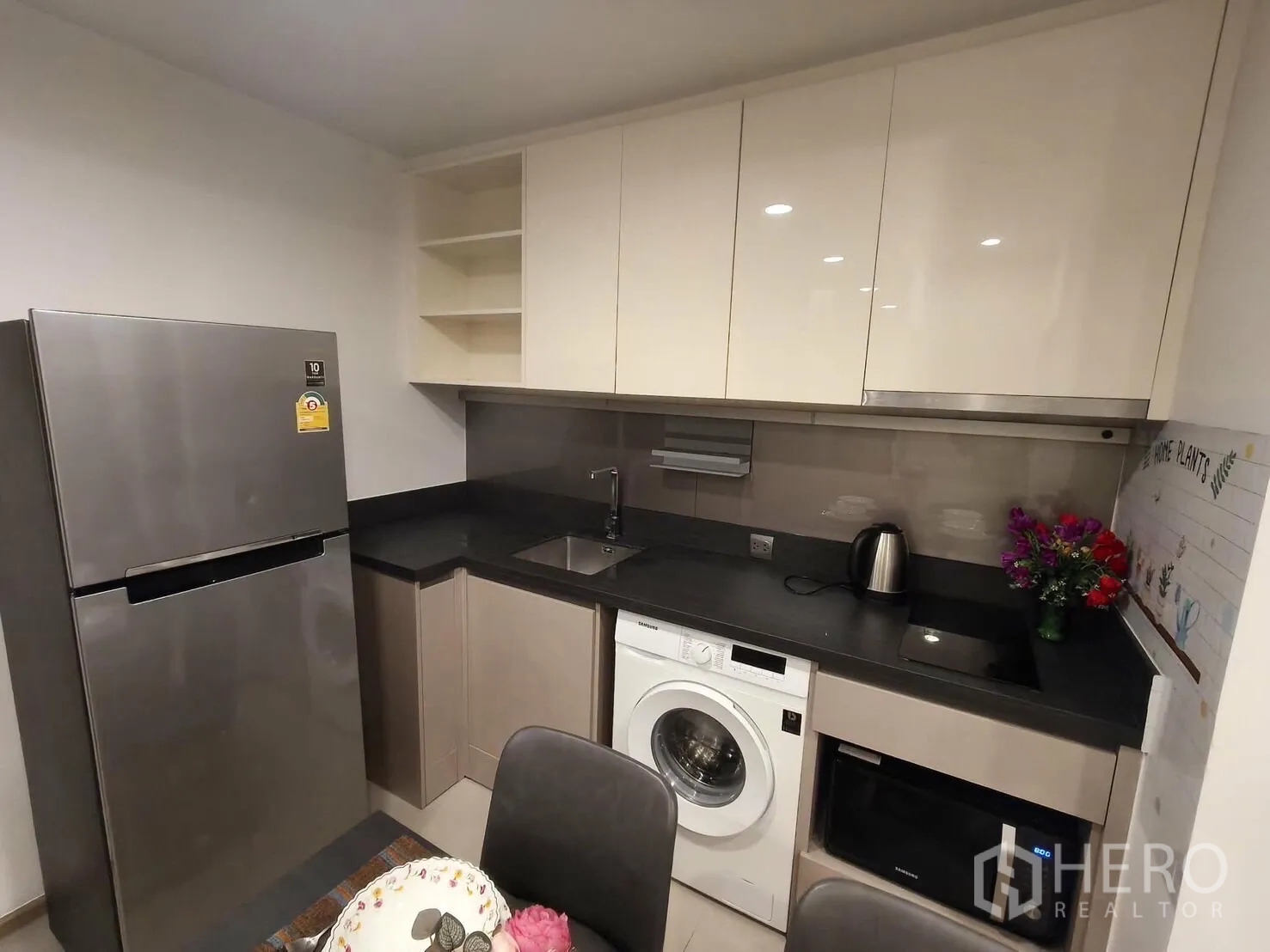 Condominium for rent in Khlong Toei, Bangkok - Compact kitchen with glossy cabinets, sink, cooktop, washing machine, microwave, and a stainless-steel fridge.