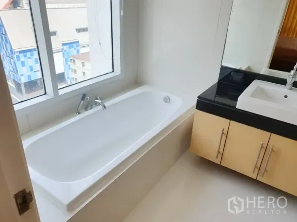 Condominium for rent or sale in Watthana, Bangkok - Bathroom with window-side bathtub and vanity with black countertop and square sink.