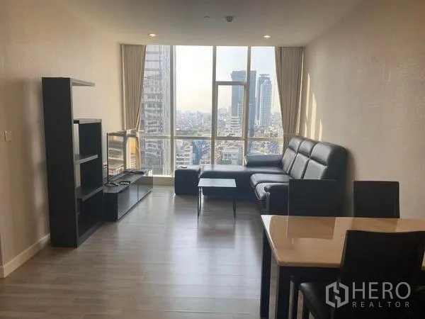 Condominium for rent or sale in Bang Rak, Bangkok - Living room with black L-shaped sofa, shelving and large windows facing Bangkok skyline.