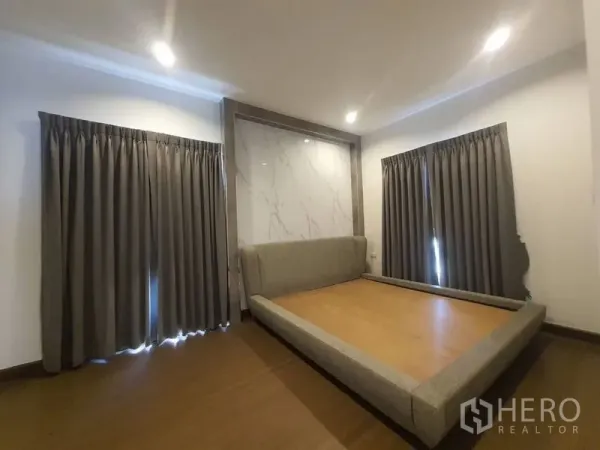 独立式住宅 出租在Prawet, Bangkok - 卧室配平台床、仿大理石背景墙、木地板与深色窗帘。