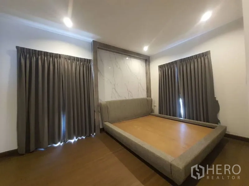 独立式住宅 出租在Prawet, Bangkok - 卧室配平台床、仿大理石背景墙、木地板与深色窗帘。