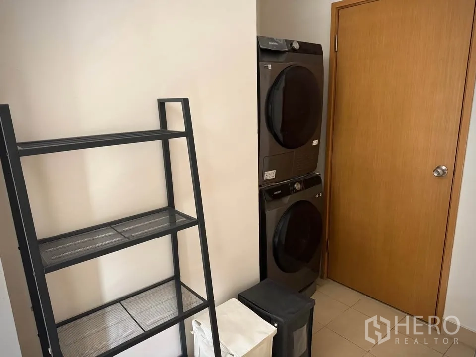 Condominium for rent or sale in Khlong Toei, Bangkok - Utility room with stacked washer and dryer beside metal shelving and a wooden door.
