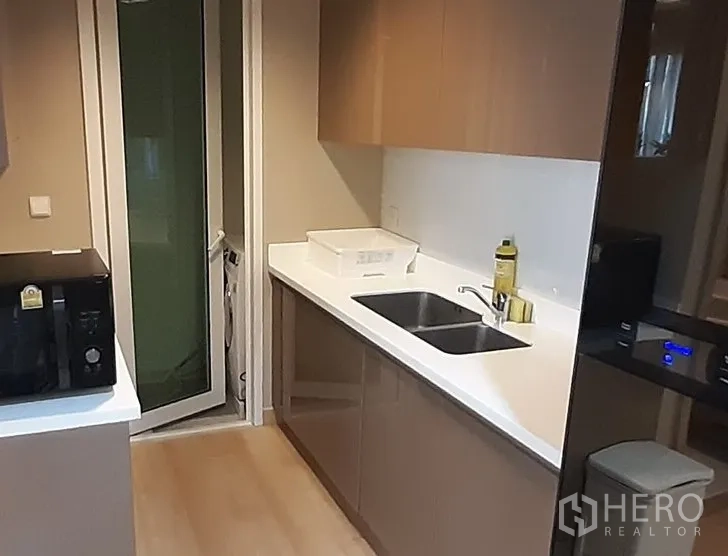Condominium for rent in Khlong Toei, Bangkok - Sleek galley kitchen with white countertops, double sink, upper cabinets and microwave.