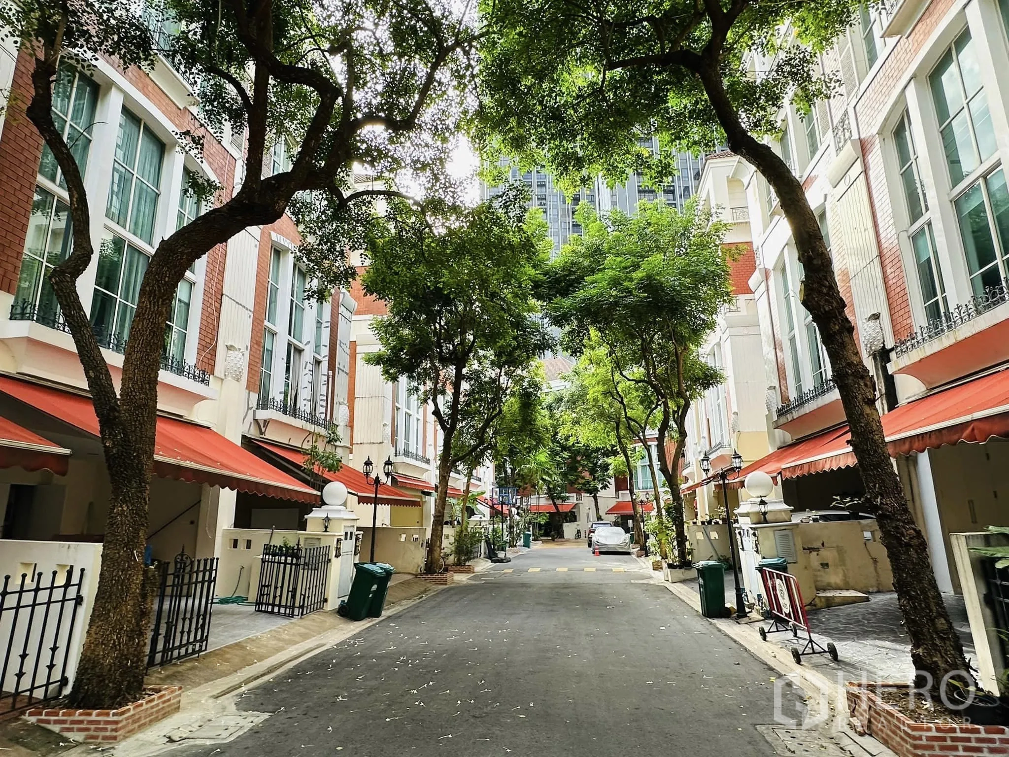 联排别墅 出租在Watthana, Bangkok - 社区内部林荫道路，两侧联排住宅配橙色遮阳篷与入户小门