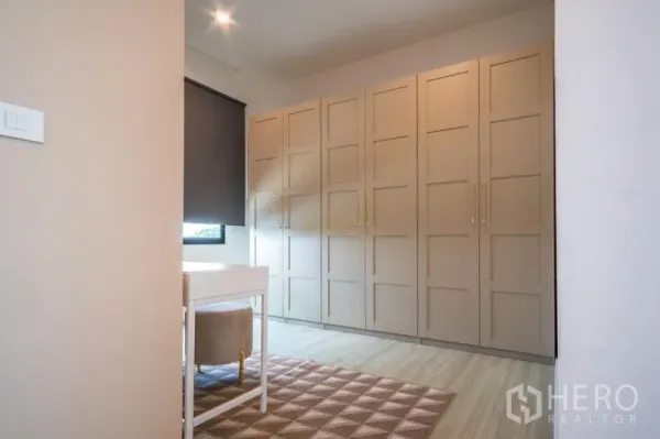 Townhouse for rent or sale in Suan Luang, Bangkok - Dressing room with built-in beige wardrobes, a vanity desk, and a window with blackout blind.