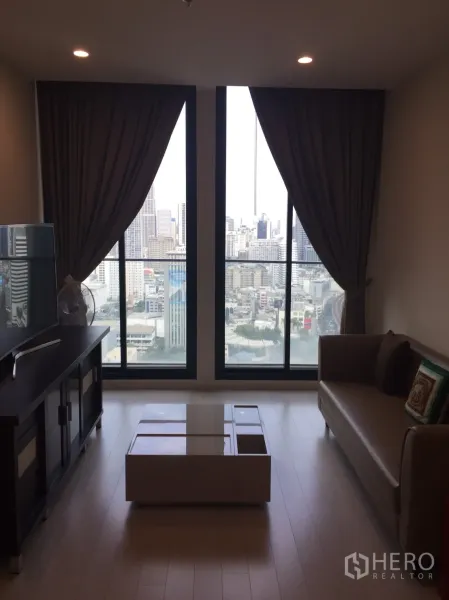 Condominium for rent in Pathum Wan, Bangkok - Living room with floor-to-ceiling windows, city skyline view, sofa, and coffee table.
