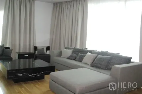 Condominium for sale in Khlong Toei, Bangkok - Close-up of L-shaped grey sofa with cushions and a black marble coffee table beside full-height curtains.
