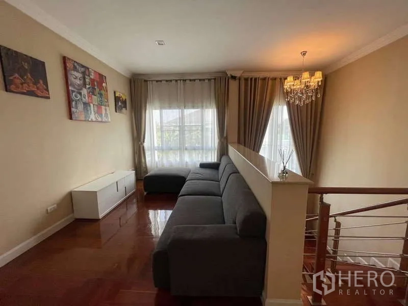 独立式住宅 出租在Mueang Nonthaburi, Nonthaburi - 二楼起居区，超长沙发、木地板与吊灯，紧邻楼梯。