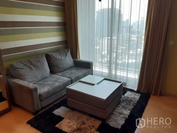Condominium for rent in Khlong Toei, Bangkok - Bright living room with gray sofa, striped feature wall, rug and floor‑to‑ceiling window to a balcony city view.