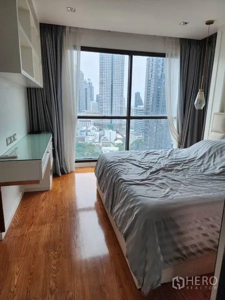 Condominium for rent in Bang Rak, Bangkok - Bedroom corner with floor‑to‑ceiling window, city view, built‑in desk and grey drapes.