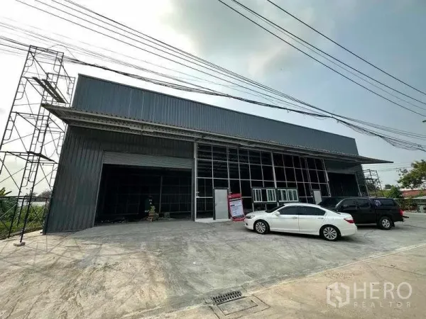 货栈 出租在Pak Kret, Nonthaburi - 新建金属板外墙仓库外观，卷闸门、玻璃办公区与宽阔停车区域（Pak Kret）。