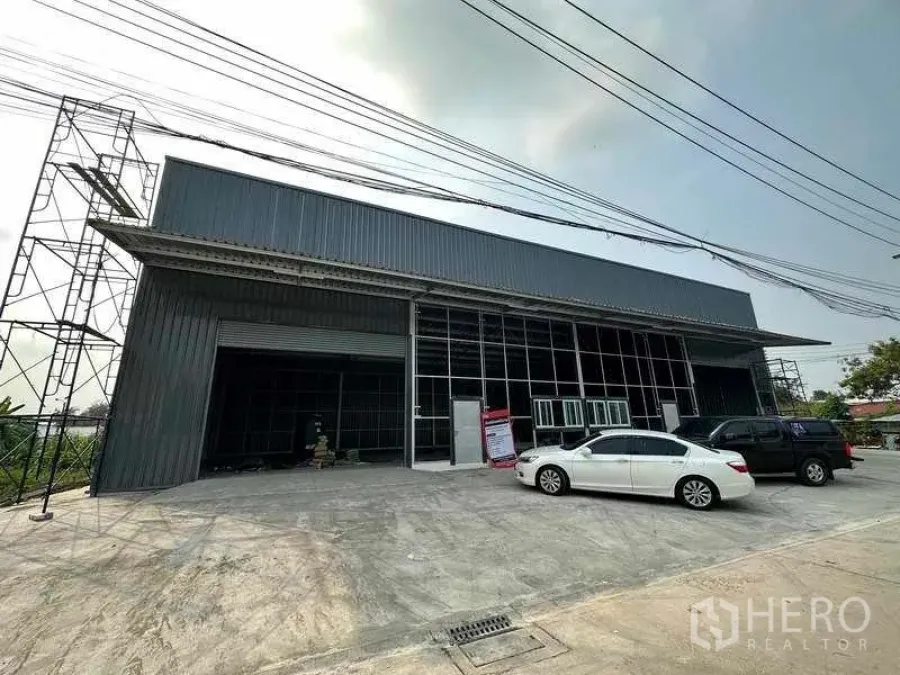 货栈 出租在Pak Kret, Nonthaburi - 新建金属板外墙仓库外观，卷闸门、玻璃办公区与宽阔停车区域（Pak Kret）。