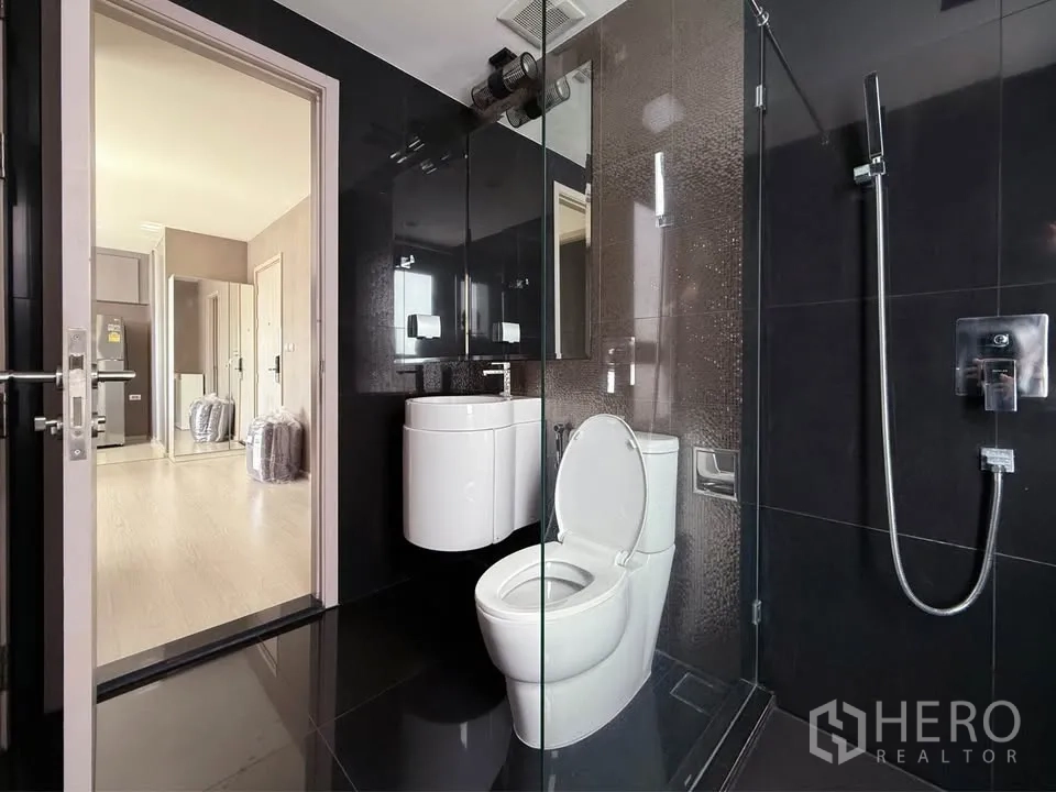 公寓 for rent in Khlong Toei, Bangkok - 黑色瓷砖浴室，带玻璃淋浴、马桶与弧形洗手台，门外通向走廊。
